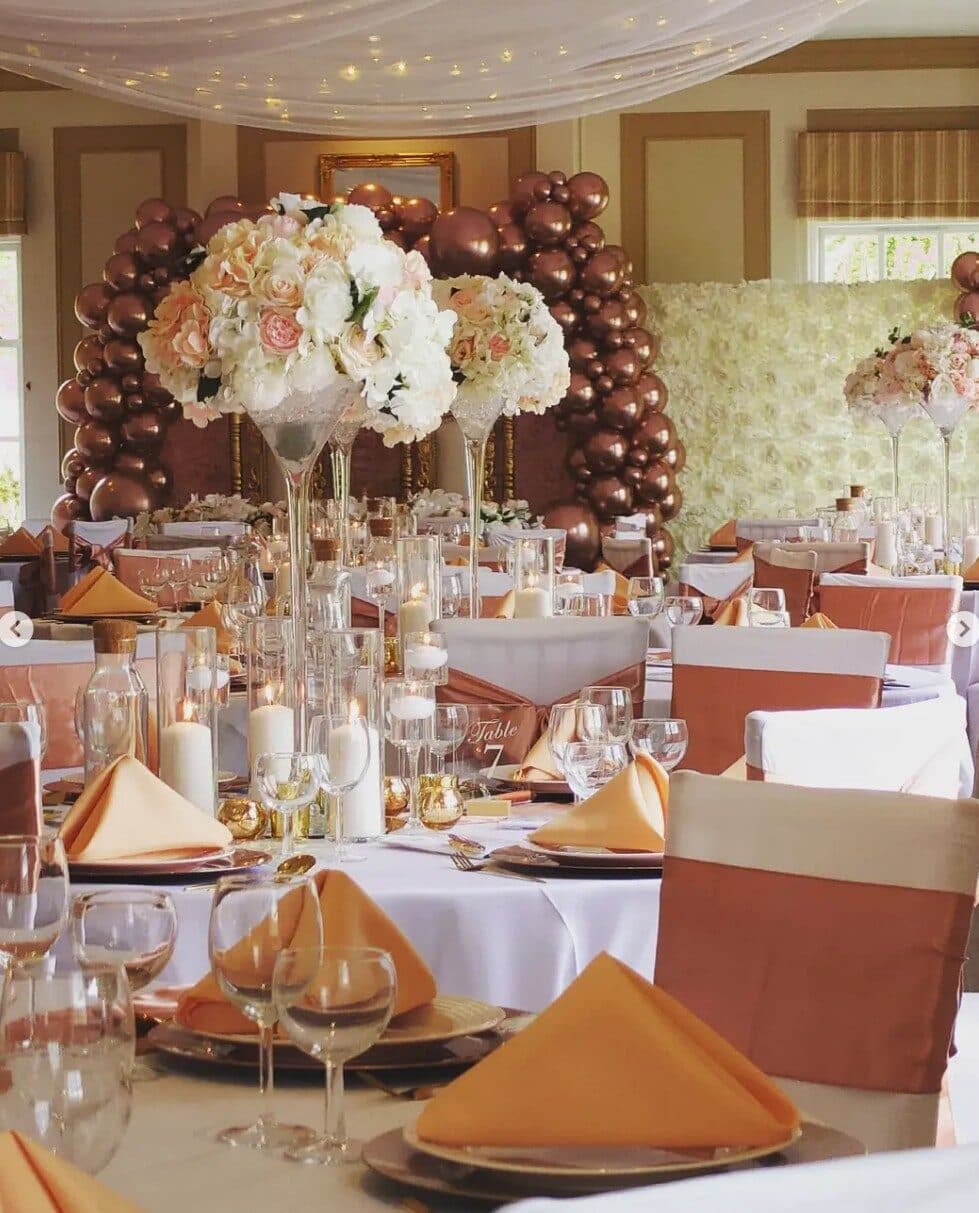 Rose gold table setting with copper napkins, a balloon garland and flower wall behind tall vase centrepieces